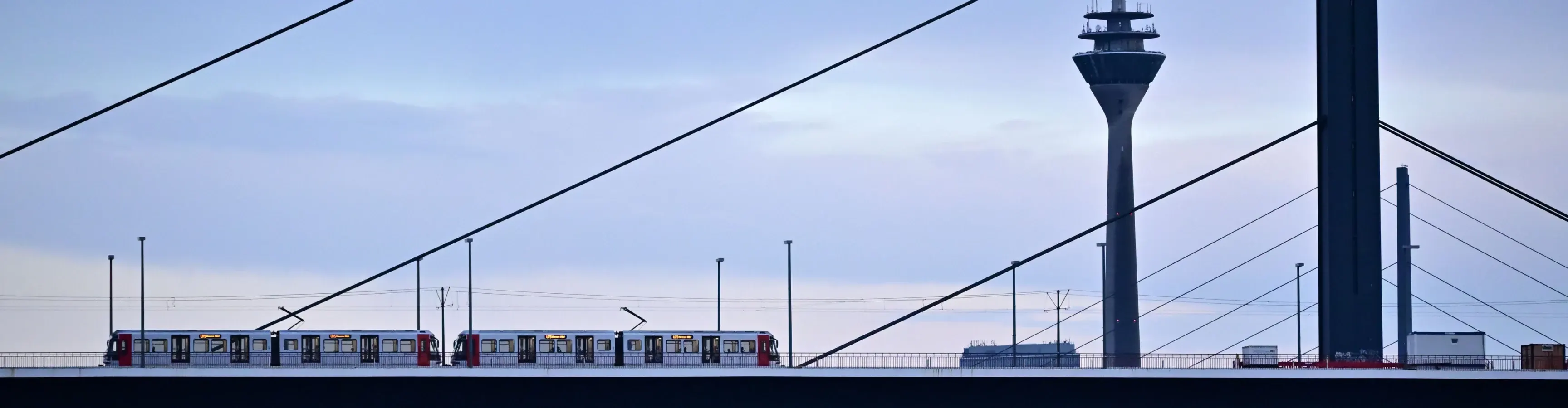 Schrägseilbrücke über den Rhein in Düsseldorf in der Abenddämmerung, dahinter der Rheinturm. Eine Stadtbahn fährt über die Brücke.